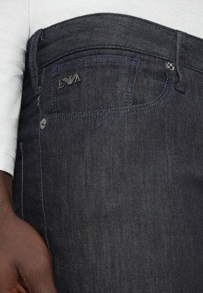 Σκούρα τζιν από ντένιμ με λεία υφή, διακοσμημένα με ένα διακριτικό λογότυπο, ραφές σε αντίθεση και μεταλλικό κουμπί στη μέση.