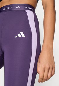 Leggings deportivos morados con una franja lateral en morado y lila, que presentan un logo de Adidas en blanco y una cintura ajustada etiquetada como "TECHFIT".