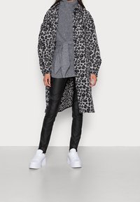 Lang leopardprint jas met knopen aan de voorkant, gecombineerd met een grijze coltrui, zwarte leren broeken en witte platform sneakers.