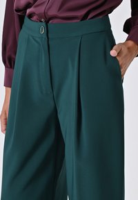 Pantaloni a vita alta, plissettati in tessuto verde scuro, con chiusura a bottone e tasche laterali. Texture liscia con vestibilità su misura.