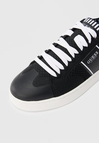 Μαύρο αθλητικό παπούτσι με λευκά κορδόνια, λευκή σόλα, ύφασμα από δίχτυ και λογότυπο "Guess Los Angeles" στην πλευρική επιφάνεια.