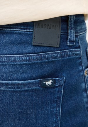 Jeans in denim blu scuro con una toppa in pelle etichettata "MUSTANG" e un piccolo logo di cavallo bianco sulla tasca. Caratteristiche includono cuciture delicate e una vestibilità slim.