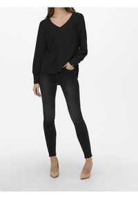 Schwarze, langärmelige Bluse mit V-Ausschnitt und strukturiertem Stoff, kombiniert mit dunklen Skinny Jeans und beigen High Heels.