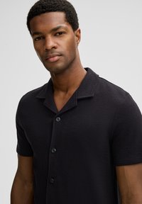 Junger Mann mit kurzem, lockigem Haar, der ein schwarzes, kurzärmliges, geknöpftes Hemd mit Kragen trägt und vor einem schlichten, hellen Hintergrund steht.