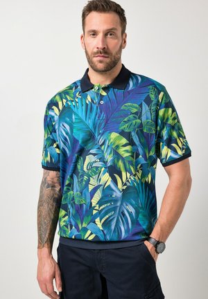 Mann mit kurzen Haaren und Bart, trägt ein marineblaues Polohemd mit kurzen Ärmeln und lebhaftem blau-grünem tropischem Blattmuster, marineblaue Hose und eine Armbanduhr.