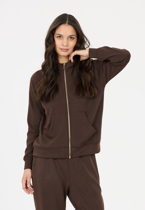 Woman with long dark hair wearing a brown zip-up jacket and matching pants, posing with one hand near her face against a plain background.