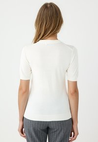 Weißes kurzärmliges Strickoberteil mit rundem Ausschnitt und geripptem Saum. Weiche Textur und leichtes Material. Getragen mit grauen Nadelstreifenhosen.