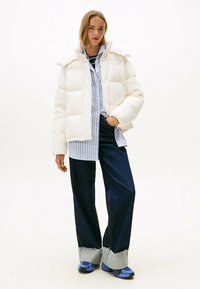 Chaqueta de plumas blanca con cuello alto, diseño acolchado y cierre a presión, sobre una camisa de botones a rayas azules y pantalones de mezclilla oscuros.