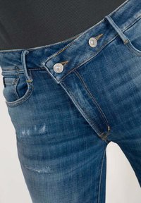 Jean en denim bleu délavé avec une fermeture à boutons, deux poches avant et des coutures oranges contrastantes. Présente un léger signe d'usure.