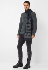 Marineblauer Schal mit auffälligem "Brax"-Logo, um den Hals drapiert. Grauer Strukturpullover und dunkle marineblaue Hose. Schwarze Schnürstiefel mit dicken Sohlen.