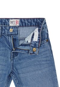 Jean en denim bleu avec braguette à fermeture éclair, bouton en métal doré et accents de couture rouge. L'étiquette indique la marque et la taille. Poche intérieure visible.