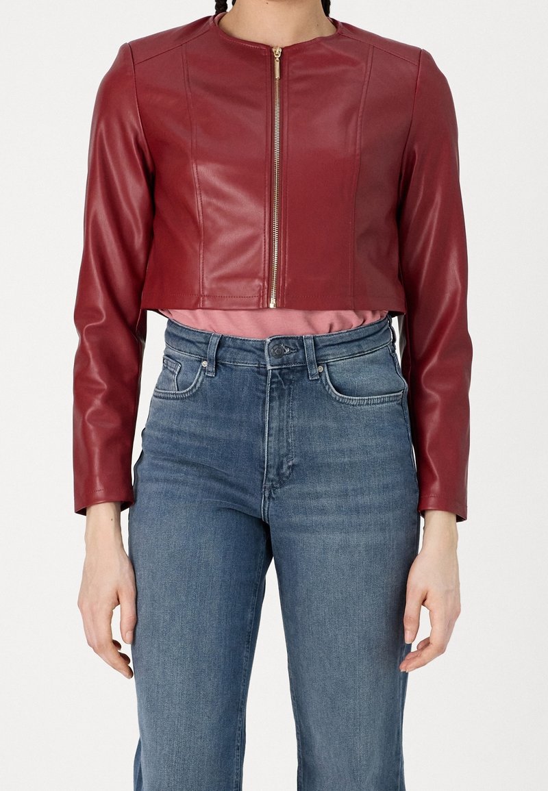 Femme portant une veste en cuir rouge courte avec une fermeture éclair à l'avant, un haut rose et un jean bleu taille haute, debout devant un fond uni.