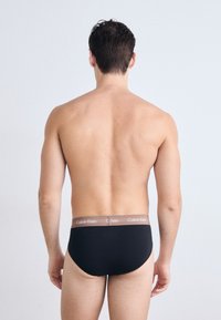 Zwarte katoenen slips met een bruine Calvin Klein-logo tailleband, strakke pasvorm, gladde textuur en een lage taille. Zicht van de achterkant.