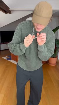 Grüner, strukturierter Pullover, dunkle Hose und eine hellbraune Mütze. Hält eine silberne Kettenhalskette. Innenraum mit Holzboden und Pflanzen.