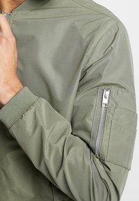 Olivenfarbener Bomberjacke mit einem Reißverschlussseitenfach am Arm, gerippten Bündchen und einem glatten, leichten Stoff mit matter Oberfläche.