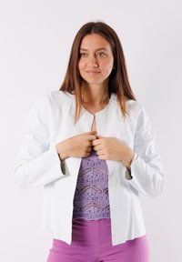 Giacca di pelle bianca con scollo rotondo e senza chiusura, caratterizzata da una texture liscia e un design minimalista. Indossata sopra un top in pizzo viola.