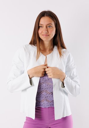 Giacca di pelle bianca con scollo rotondo e senza chiusura, caratterizzata da una texture liscia e un design minimalista. Indossata sopra un top in pizzo viola.