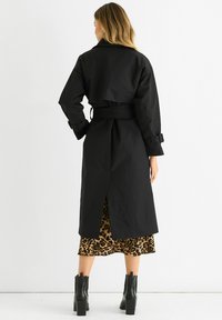Black trench coat with wide collar, belted waist, and cuffed sleeves; features a split back revealing a leopard print dress underneath.