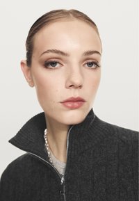 Donkergrijze ritstrui met een hoge kraag en ribbelstructuur, gedragen over een lichtgrijze shirt. Model heeft minimale makeup en natuurlijk haar.