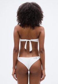 Λευκό μπικίνι με τοπ μπαντό που έχει δετή λεπτομέρεια στην πλάτη. Τα σλιπ είναι χαμηλού κόψιμου με λογότυπο στη αριστερή πλευρά. Λείο υφασμάτινο υλικό.