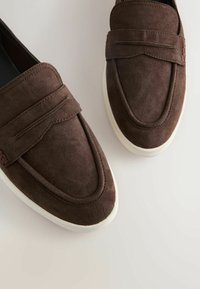 Mocassins en daim brun foncé avec semelles en caoutchouc blanches, présentant une bande décorative sur le dessus, exposés sur un fond clair.