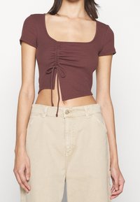 Femme portant un crop top marron à manches courtes avec un cordon à l'avant et un pantalon beige taille haute.