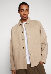 Veste en coton beige avec un devant à boutons, deux grandes poches et des coutures blanches. Portée sur un t-shirt blanc et un pantalon vert.