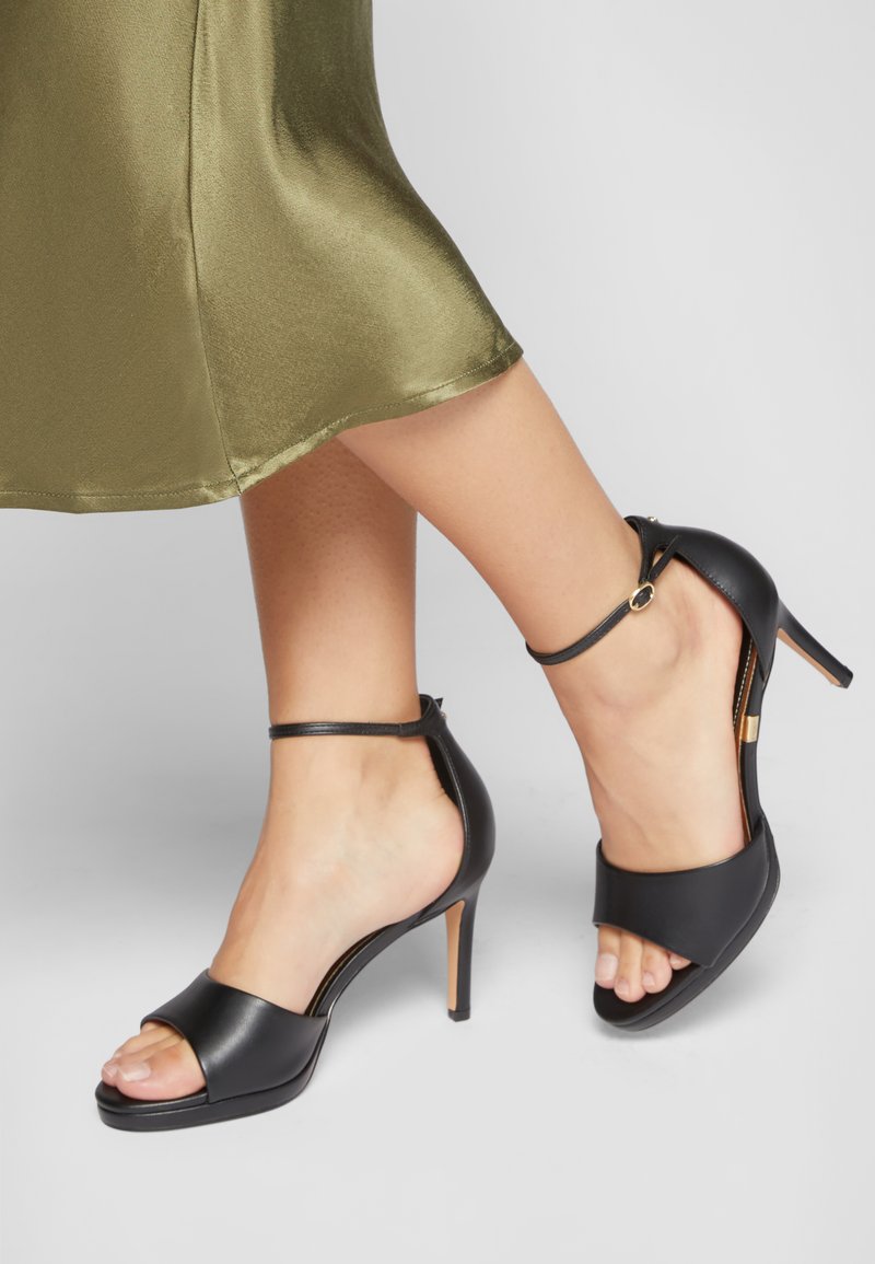 Sorte læder højhælede sandaler med åben tå, ankelrem og et slankt design. Glat tekstur med en guld accent detalje bagpå.