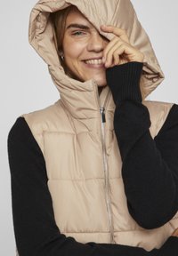Gilet en duvet beige avec capuche et fermeture éclair sur le côté, porté sur un pull en maille noir. Le gilet présente une texture matelassée et un col haut.