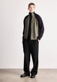 Atlétikus cipzáras dzseki olívazöld és fekete színekben, magas gallérral, kontrasztos varrással és oldalzsebekkel, laza fekete nadrággal párosítva.