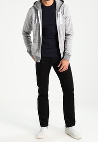 Felpa grigia con zip e cappuccio, con stringhe bianche, indossata sopra una t-shirt blu navy, abbinata a jeans neri e sneakers bianche.