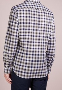 Mężczyzna w dopasowanej koszuli z długim rękawem w czarno-białą szaro-skalową kratkę, w połączeniu z ciemnymi spodniami, widok z tyłu.