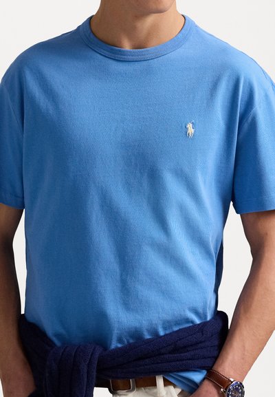 Blå kortärmad T-shirt i ett strukturerat tyg. Har en liten vit logotyp på bröstet. Bärs med en marinblå tröja knuten i midjan.