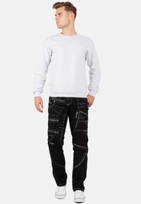 Felpa grigia e pantaloni cargo neri con cuciture bianche, caratterizzati da molteplici tasche e motivi, abbinati a sneakers bianche.