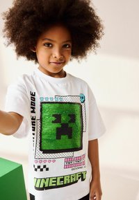 La t-shirt bianca a maniche corte presenta un volto di Creeper di Minecraft in paillettes verdi, dettagli neri e un testo grafico vicino all'orlo.