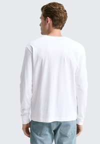 Mann, der sich abwendet und ein schlichtes weißes Langarmshirt sowie helle blaue Denim-Jeans trägt, vor einem neutralen Hintergrund.