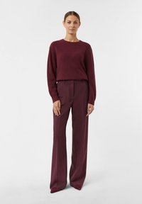 Woman stands wearing a matching burgundy knitted sweater and wide-leg trousers with pointed-toe shoes against a white background.