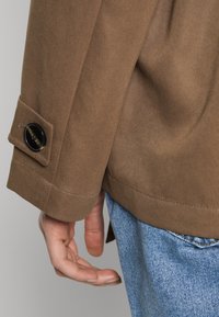 Bruine jas met een gladde textuur, voorzien van een zwart knoopdetail op de manchetten, gecombineerd met lichtblauwe spijkerbroek.
