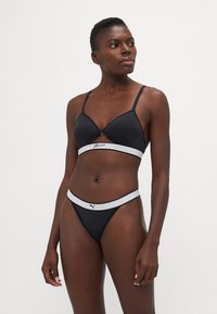 Black bra with triangular cups and adjustable straps, featuring a white band with logo. Black thong with a contrasting waistband.