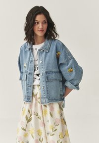 Lichtblauwe spijkerjack met een oversized pasvorm, voorkzosk pockets en geborduurde patches. Draag het over een witte graphic T-shirt en een bloemenrok.