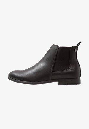 Botín Chelsea negro de cuero sin cordones con panel lateral elástico, tacón bajo y lengüeta para tirar sobre fondo blanco.
