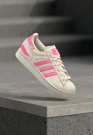 Baskets crème avec des accents roses, présentant une tige en cuir texturée, un design à trois bandes, un renfort en caoutchouc au niveau des orteils et une semelle contrastante.