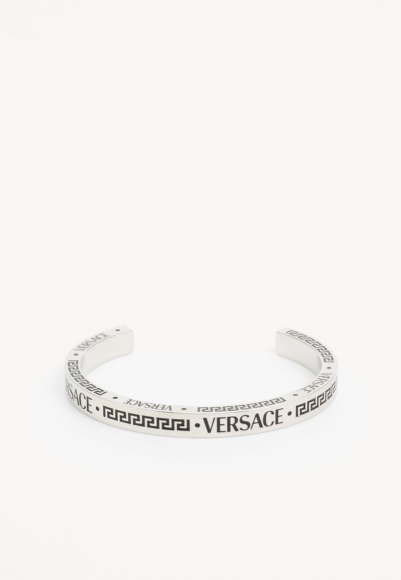 Srebrna narukvica s manžetom koja sadrži ugravirani "Versace" logo duž trake, ukrašena geometrijskim uzorkom grčkog ključa. Glatka površina.