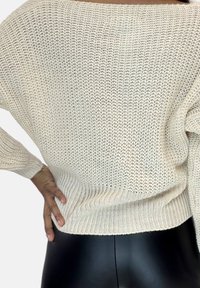 Helles, creamfarbenes Strickpullover mit lockerer Passform, weitem Ausschnitt und gerippten Bündchen. Getragen über schwarzen, dehnbaren Hosen, die den strukturierten Stoff zur Geltung bringen.