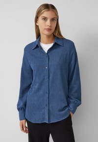 Chemise bleu texturée à col écarté, à manches longues et à coupe décontractée, portée avec une couche de base blanche en dessous.