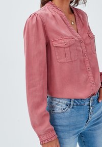 Staubrosa Bluse mit langen Ärmeln und R�üschenausschnitt, zwei Brusttaschen und Knopfverschluss, kombiniert mit hoch geschnittenen blauen Jeans.
