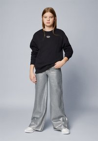 Sudadera negra de cuello redondo con un logo, combinada con unos pantalones anchos de metal plateado. Modelo luciendo zapatillas blancas, de pie frente a un fondo gris.