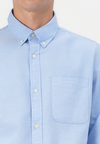 Camisa abotonada de color azul claro con cuello, que cuenta con un bolsillo en el pecho izquierdo, tejido texturizado y botones blancos a lo largo de la tapa delantera.