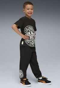 Camiseta negra con estampado gráfico blanco, combinada con pantalones de chándal negros con detalle del logotipo en la pierna. Zapatillas negras y naranjas completan el look.