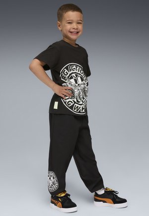 T-shirt noir avec impression graphique blanche, associé à un pantalon de survêtement noir avec un détail de logo sur la jambe. Des baskets noires et orange complètent le look.